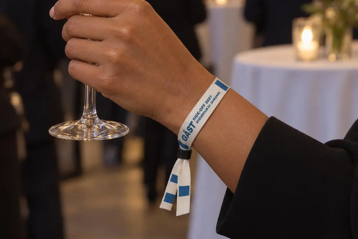 Gäst med champagneglas och ett vitt tygband med blå text, GÄST KICK-OFF 2027