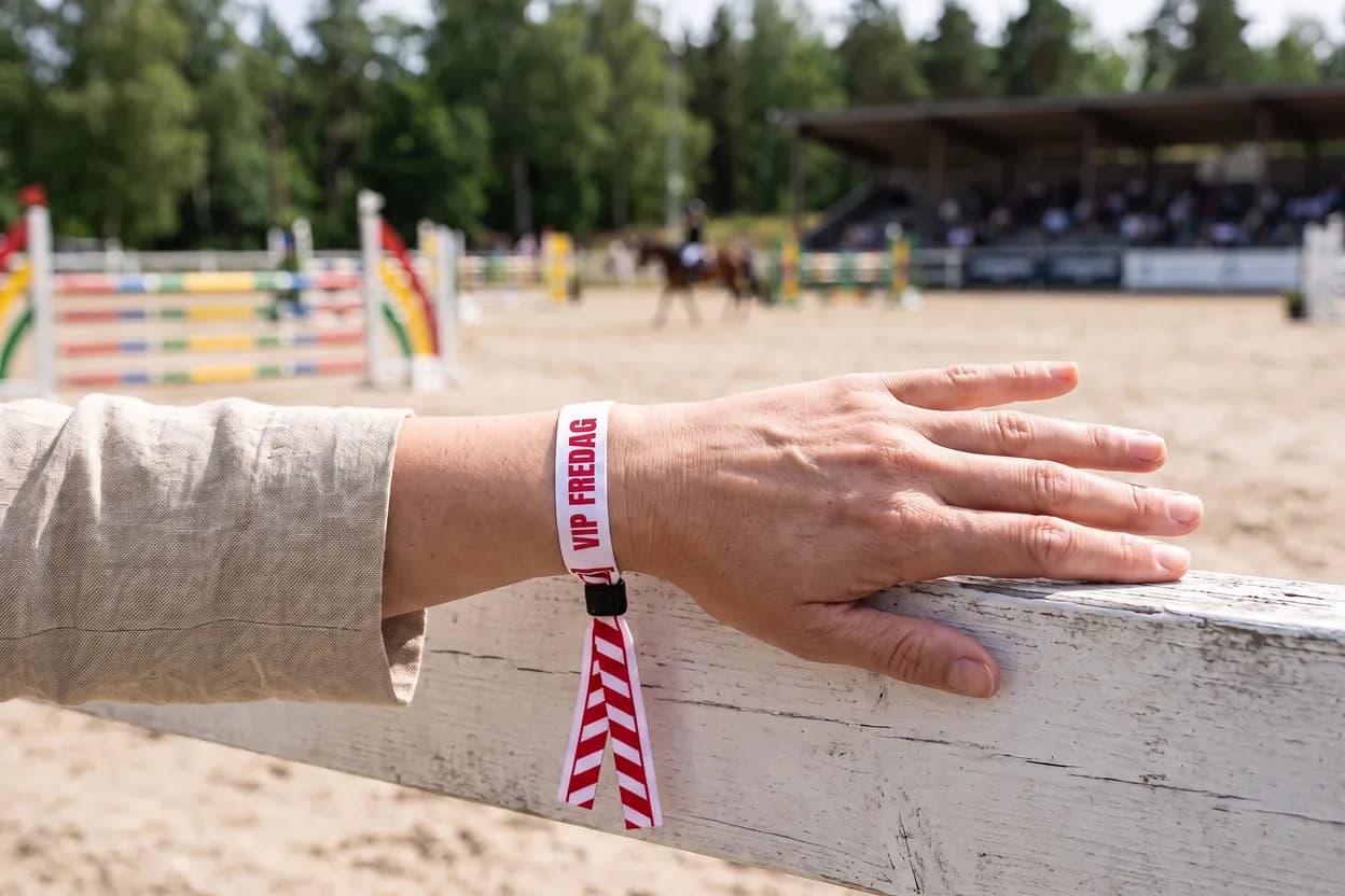 Hand am Zaun eines Springturniers mit weißem Einlassband aus Stoff, VIP FREDAG, mit rot-weiß gestreifter Hindernisstange