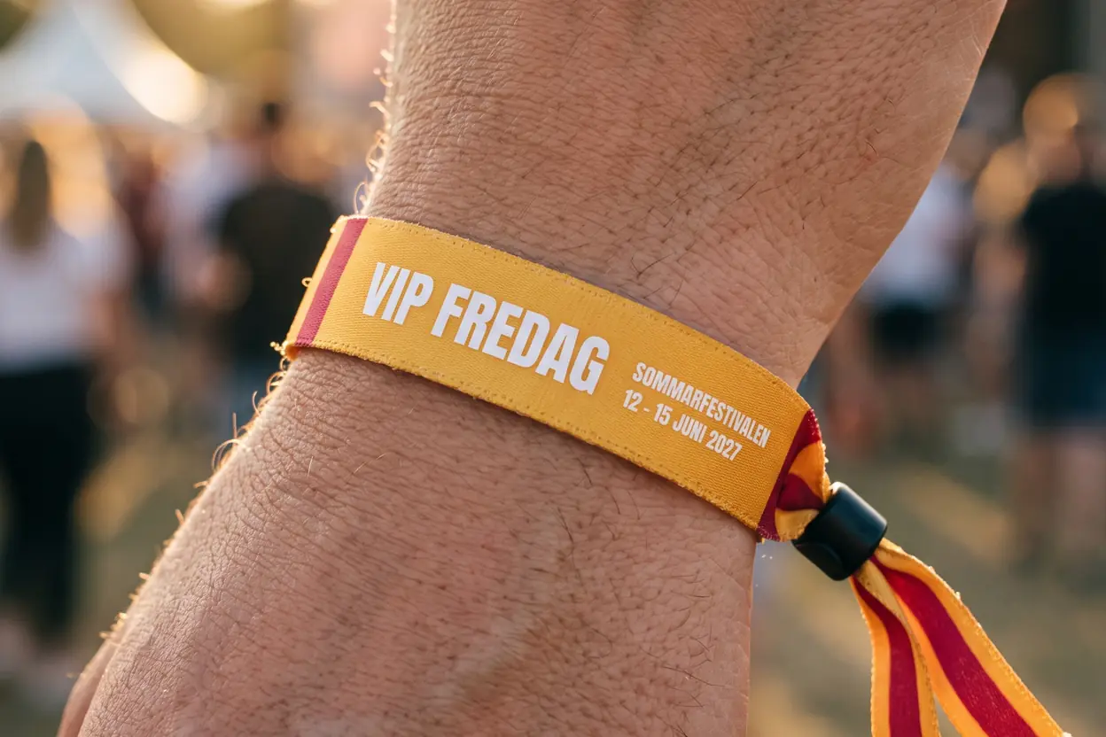 Närbild av ett gult tygband med texten VIP FREDAG, randiga ändar och svart plastlås på en handled